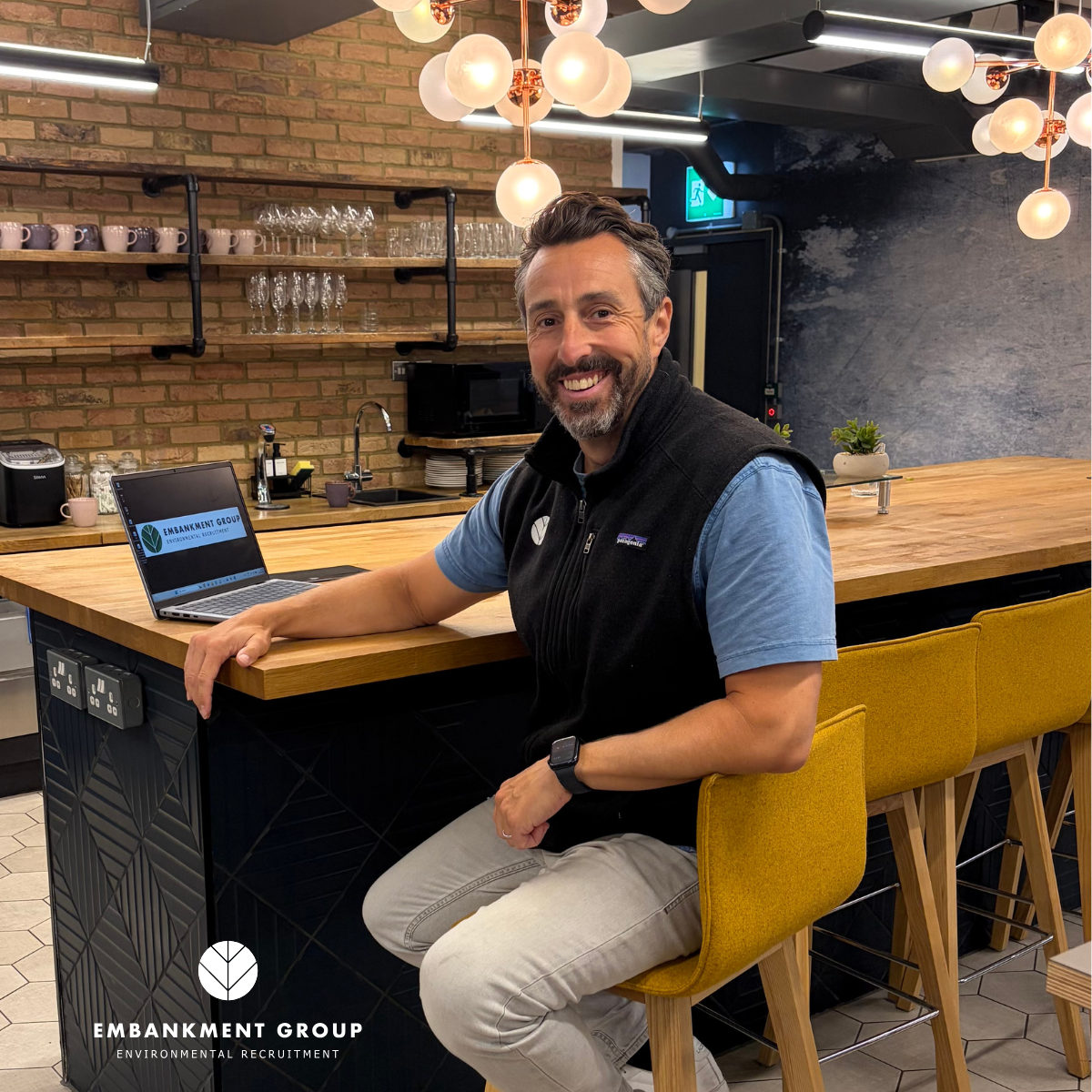 Ecology recruiter smiling at a wooden bar in an office, with hanging lights and stools.