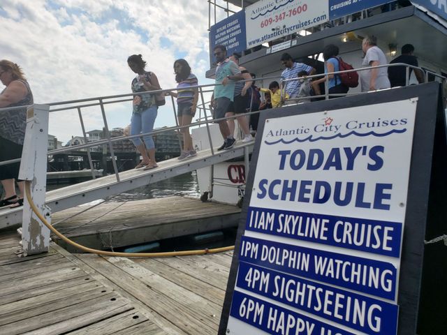 People boarding a boat; sign lists cruise schedule: Skyline, Dolphin Watching, Sightseeing, and Happy Hour.