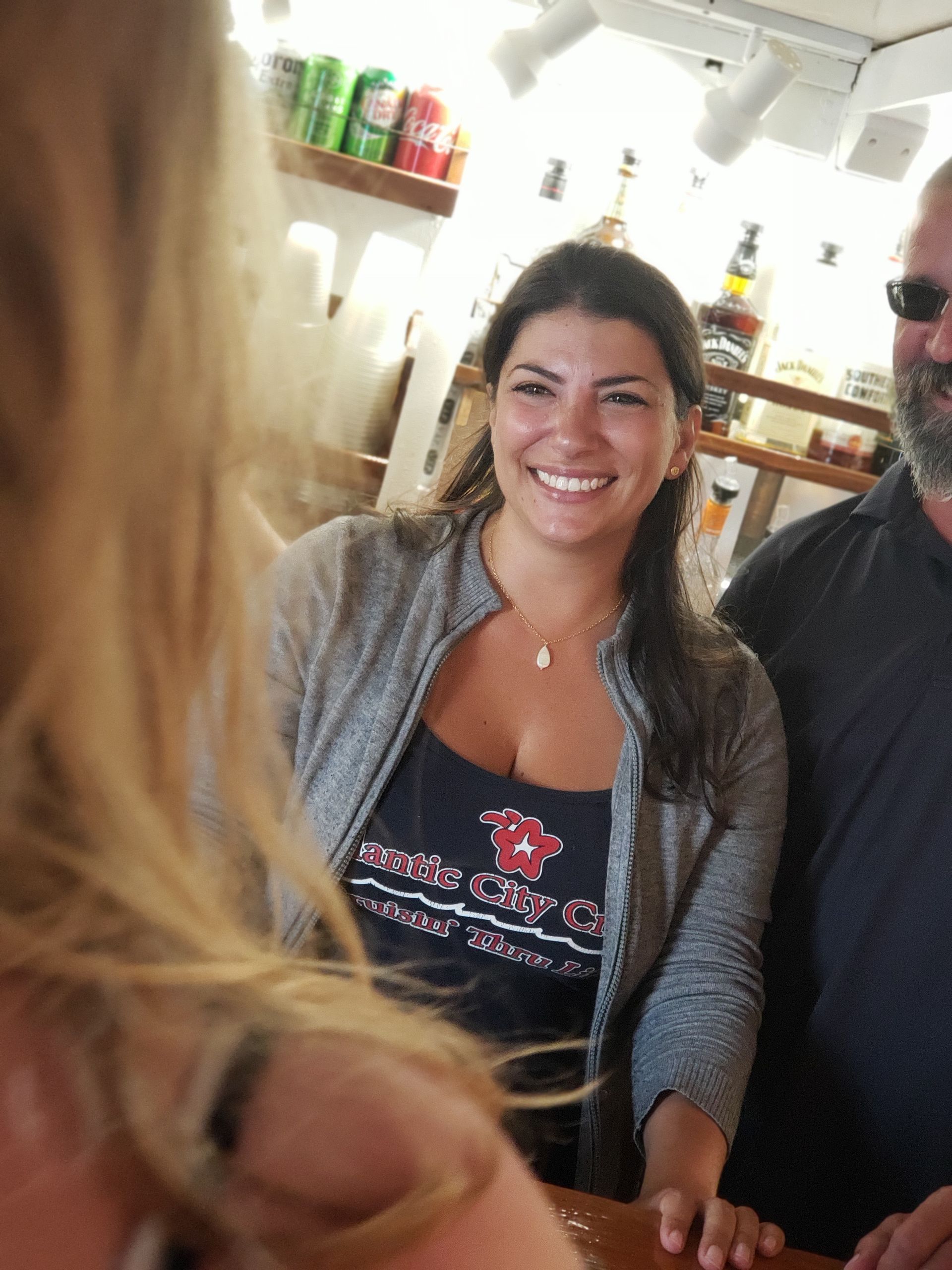 Woman smiling, wearing a gray sweater, standing at a bar.