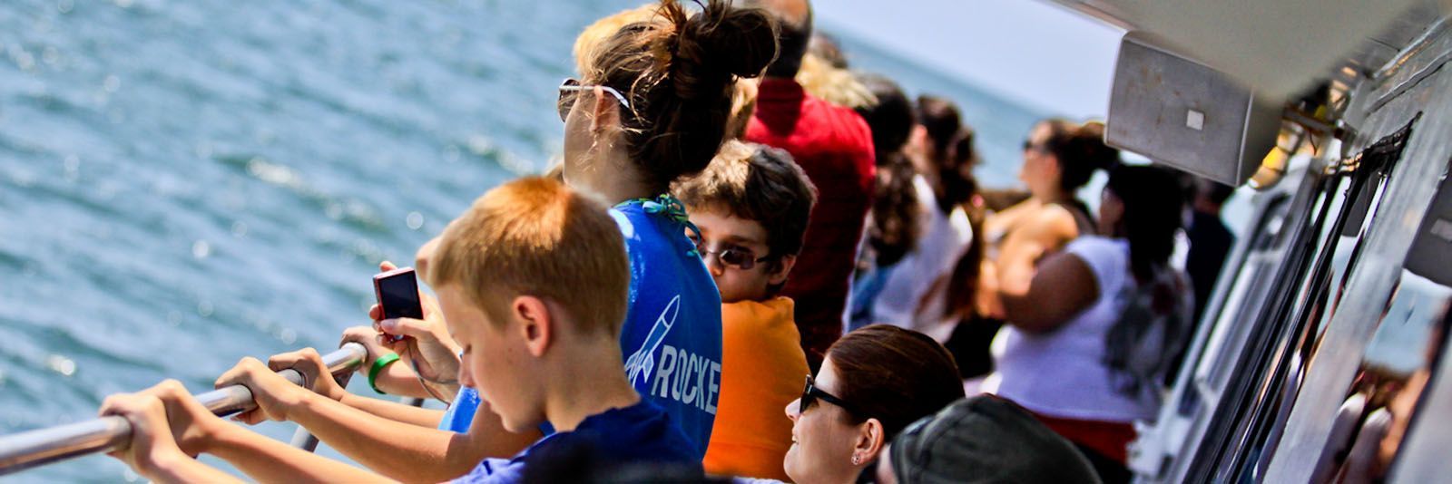 People on a boat looking at the sea. A young boy in foreground holds a phone. The sky is bright.