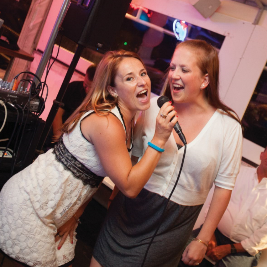 Two women singing into a microphone at an outdoor event, one wearing a white dress, the other in a sweater and skirt.