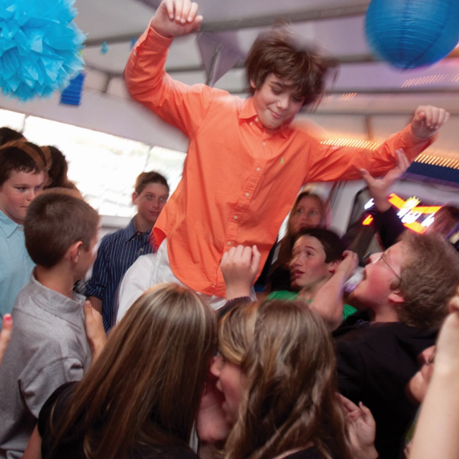 Teen boy crowd surfing at a party, orange shirt, surrounded by smiling faces, blue decorations.
