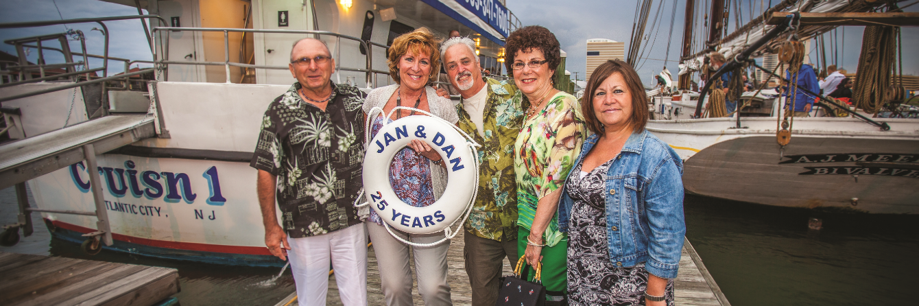 Five people pose on a dock by a boat. The boat says “Cruisn 1” and “Jersey, NJ”.  One person holds a life ring.