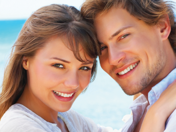Smiling couple, embracing, near the ocean. Woman on left, man on right, both smiling with bright eyes.
