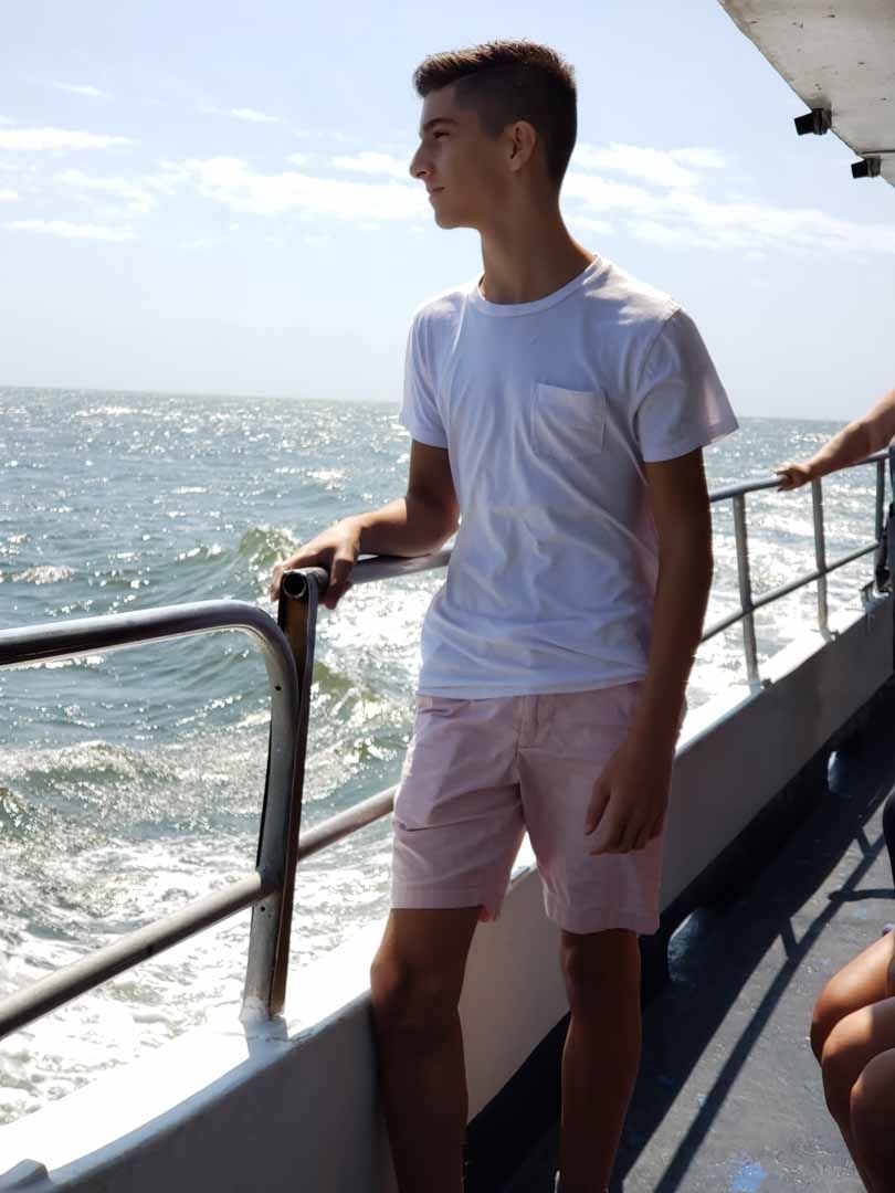 Young person on a boat, looking out at the ocean. Wearing a white shirt and pink shorts.