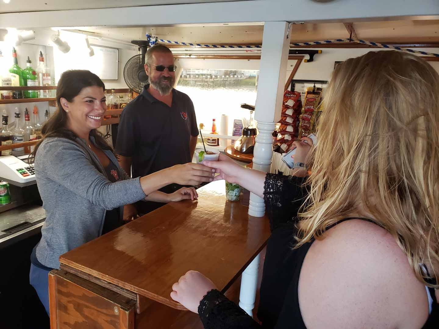 Woman handing a drink to another at a bar; bartender in the background.
