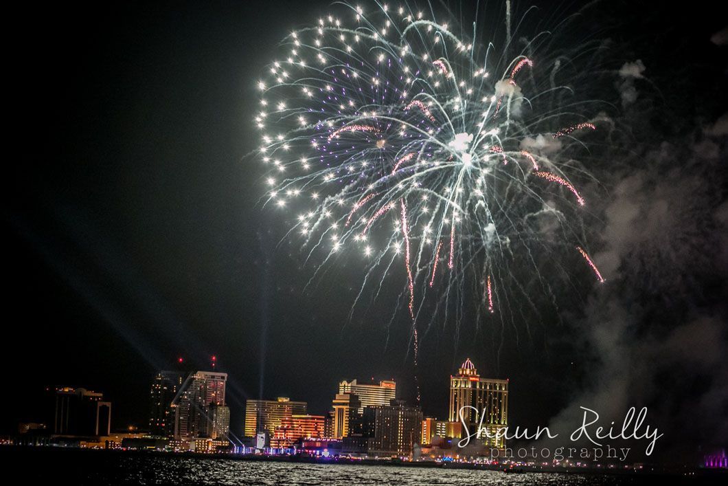 Fireworks over a city skyline at night. Bright, colorful explosions above illuminated buildings reflected in water.