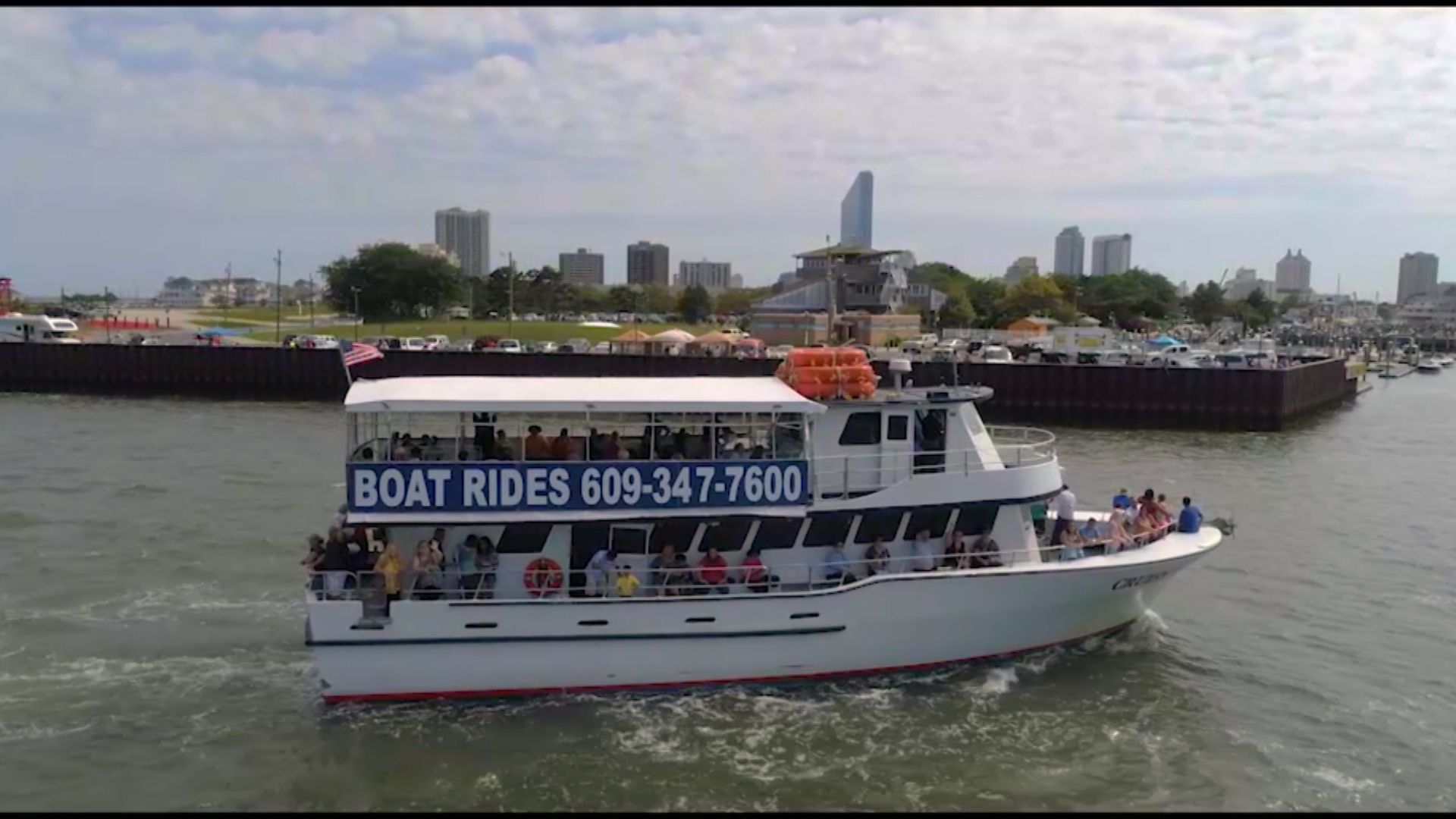 A white boat with passengers cruising on the water; a skyline is in the background. The boat says 