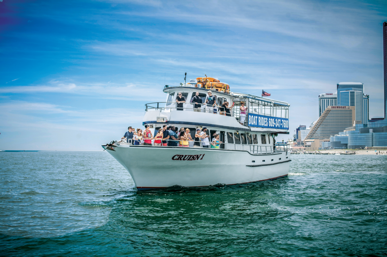 A white tour boat with passengers on the water, with a city skyline in the background under a blue sky.
