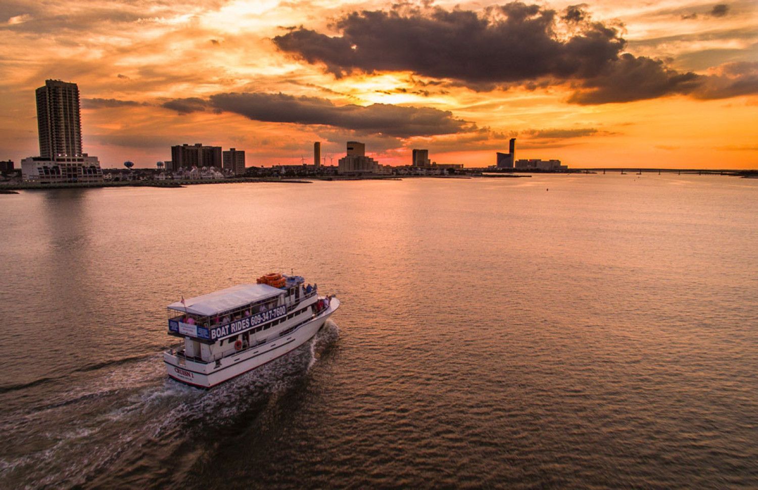 Boat sails across water at sunset, city skyline in the distance. Orange and yellow sky.