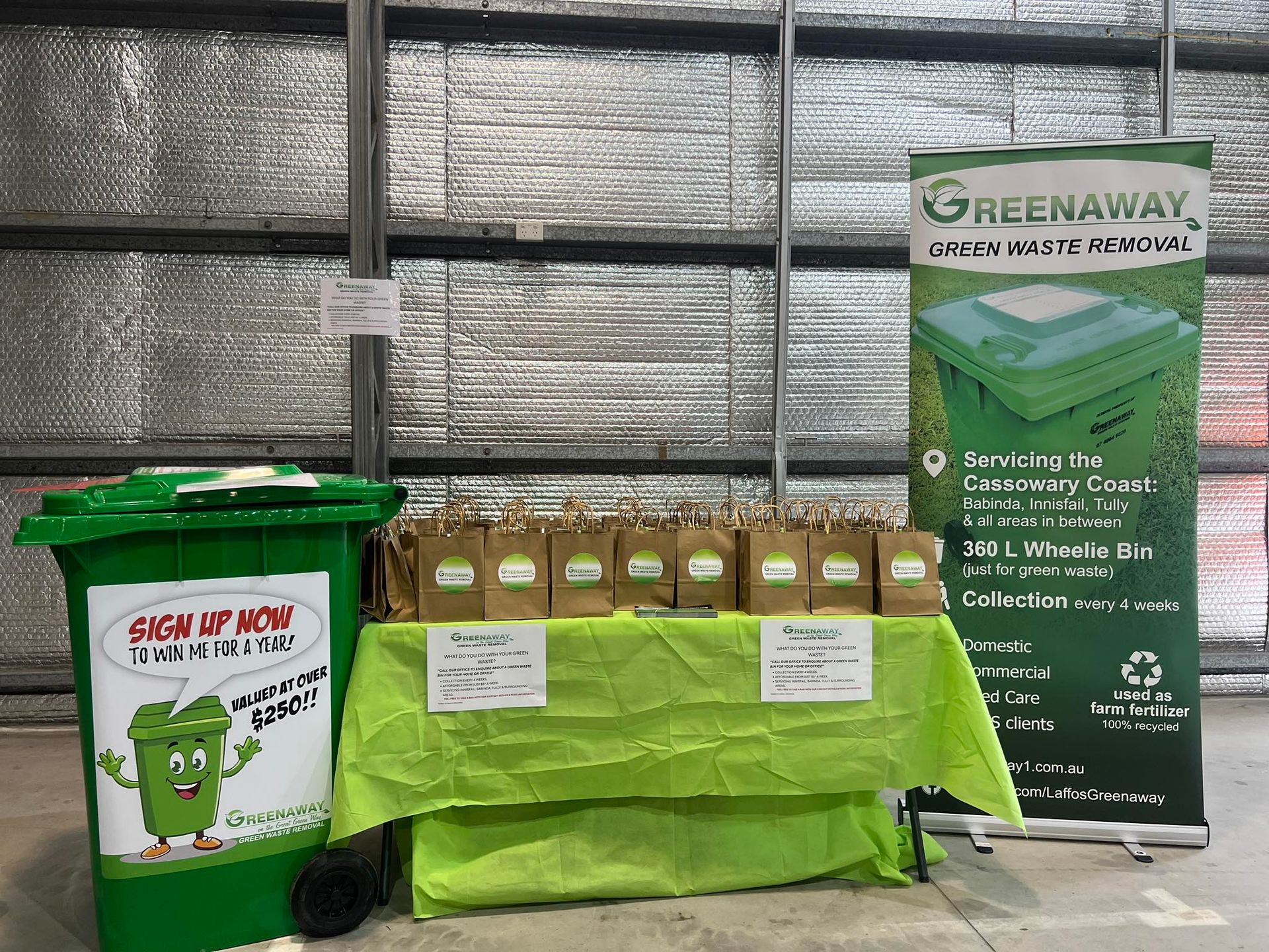 Greenaway compost bin display: a green bin, table with compostable bags, and a banner inside a warehouse.— Greenaway Green Waste Removal in O'Briens Hill, QLD