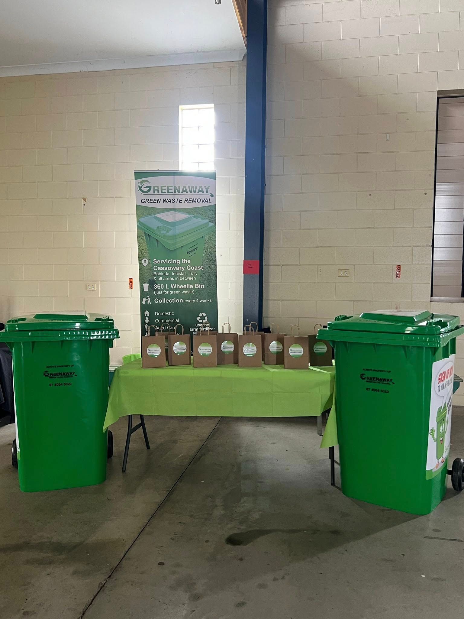 Two green bins flanking a table with brown bags and a banner in a brick-walled room.— Greenaway Green Waste Removal in O'Briens Hill, QLD