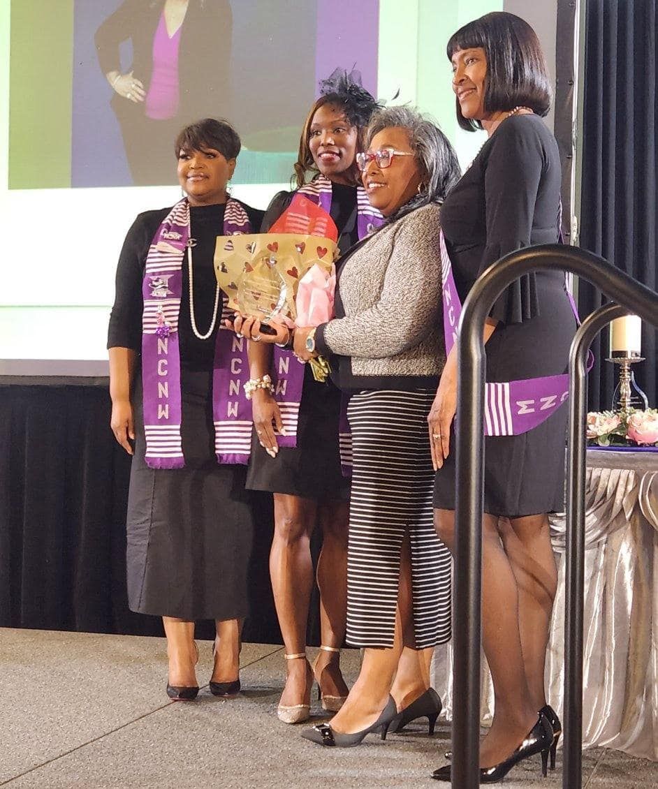 Four women in formal attire holding a gift basket on stage. Purple and black accents, celebratory setting.