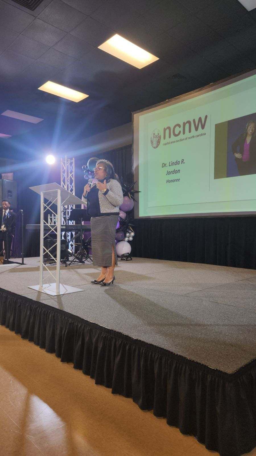 Woman speaking at podium on stage with screen displaying