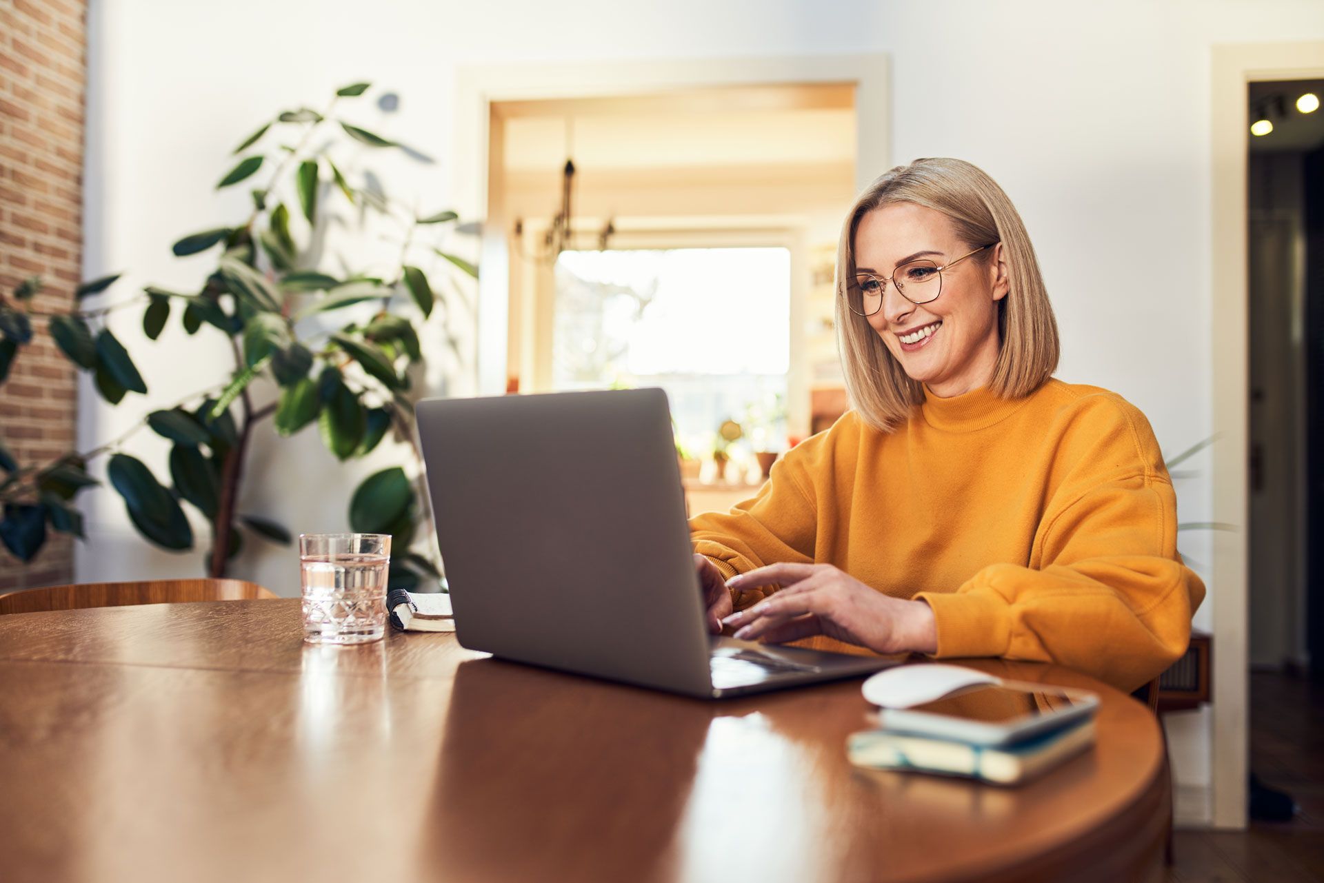 Woman with glasses, blonde hair, yellow sweater, working on laptop at a wooden table.
