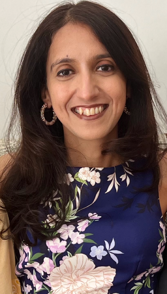 Woman with long dark hair smiles at the camera, wearing a blue floral print dress and hoop earrings.
