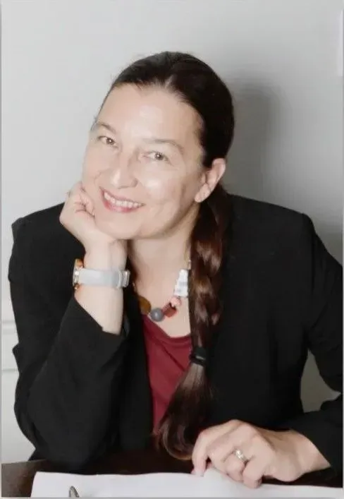 Woman with dark hair smiles, leaning on her hand, wearing a blazer and jewelry.