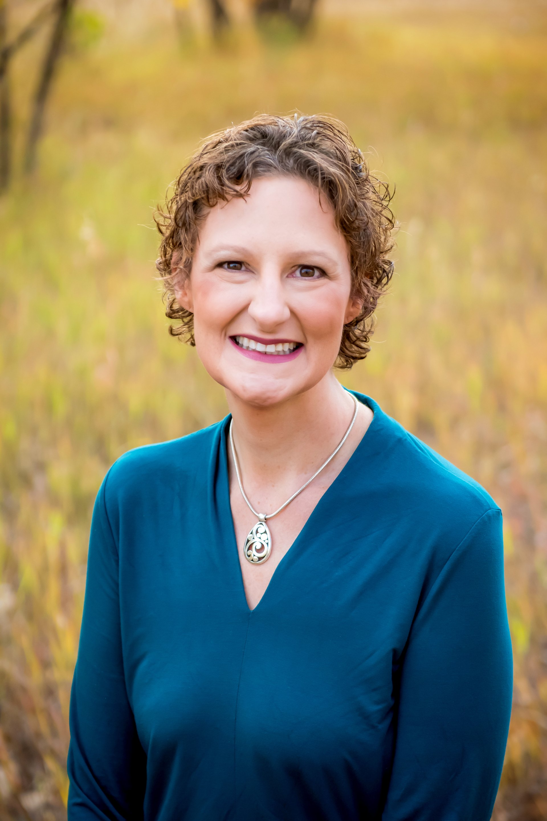 Woman with curly brown hair, smiling, wearing teal top and pendant, outdoors in field.