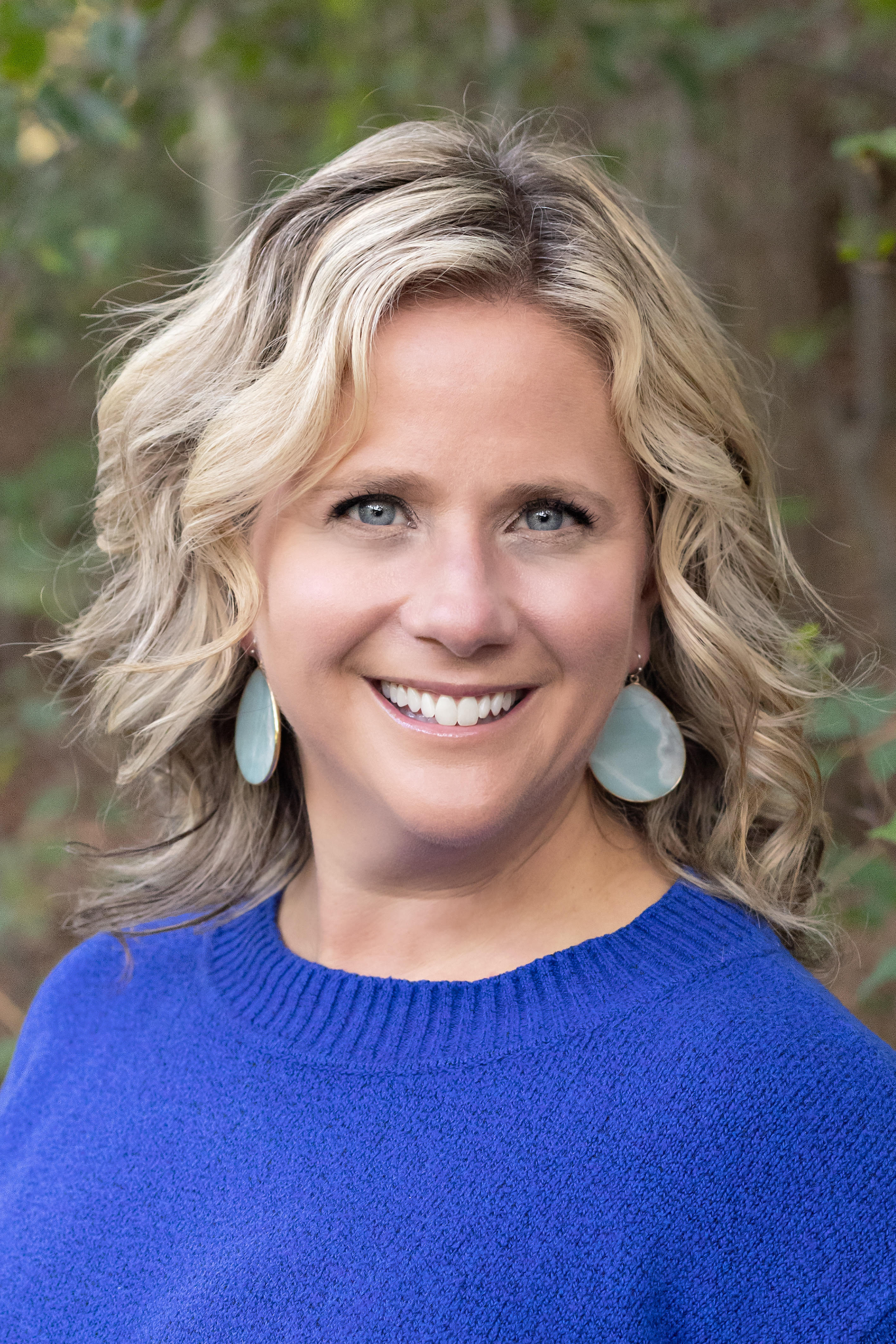 Woman with blonde hair smiles, wearing a blue sweater and turquoise earrings outdoors.