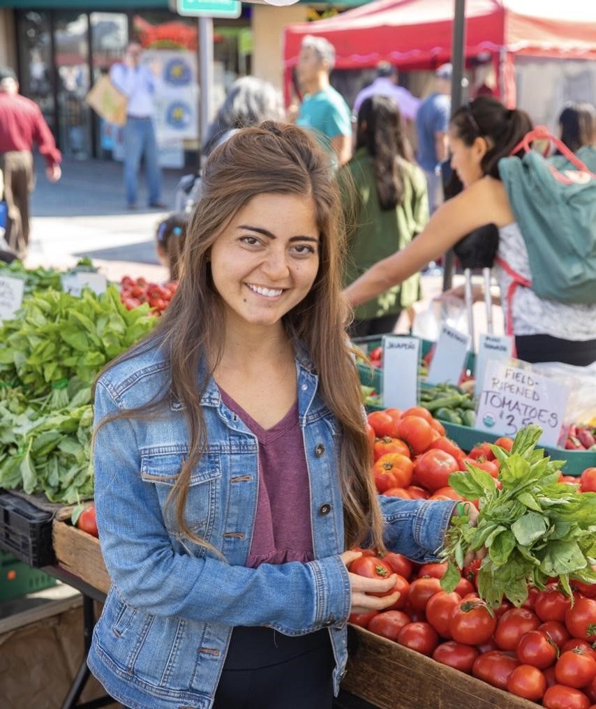 Woman smiles at farmers market, selecting a tomato from a display.