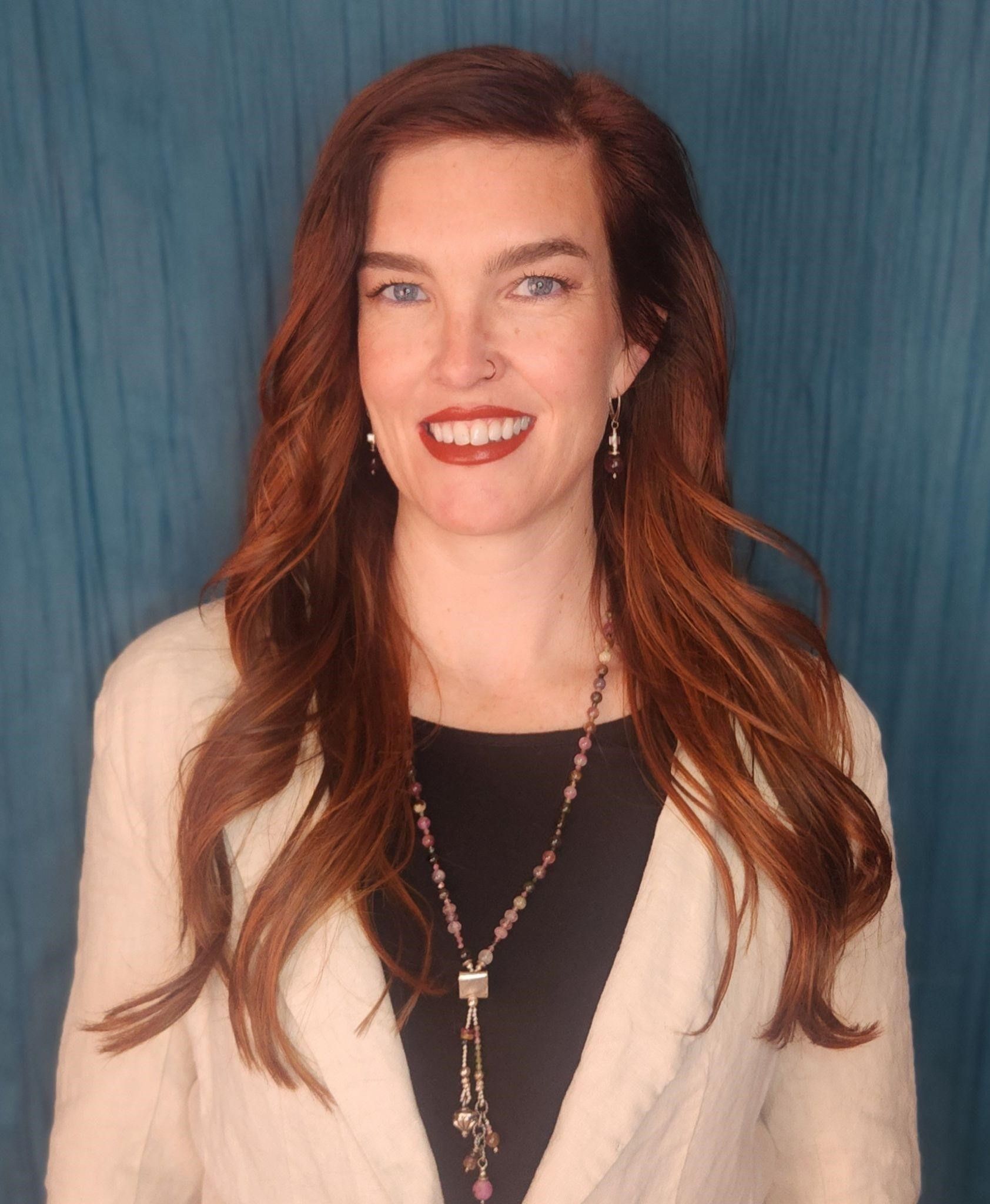 Woman with auburn hair, wearing a cream-colored sweater, black top, and beaded necklace, smiling in front of a teal backdrop.