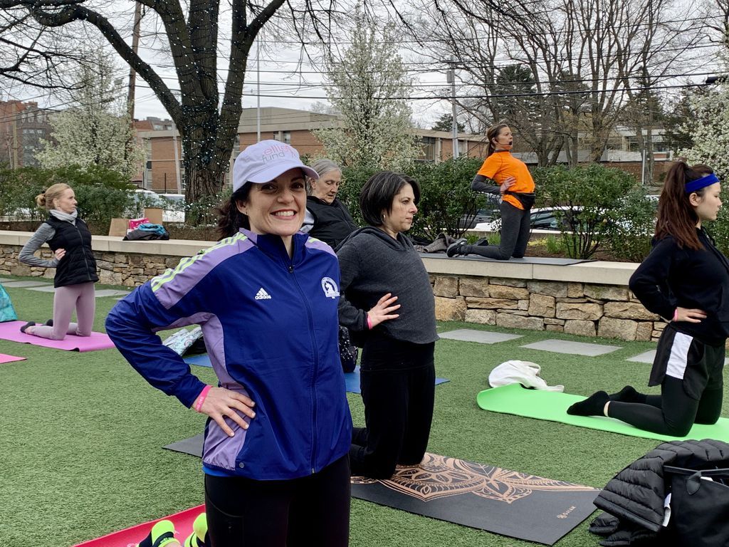 A group doing yoga outdoors; woman in blue jacket smiles, others stretch, green mats on grass.