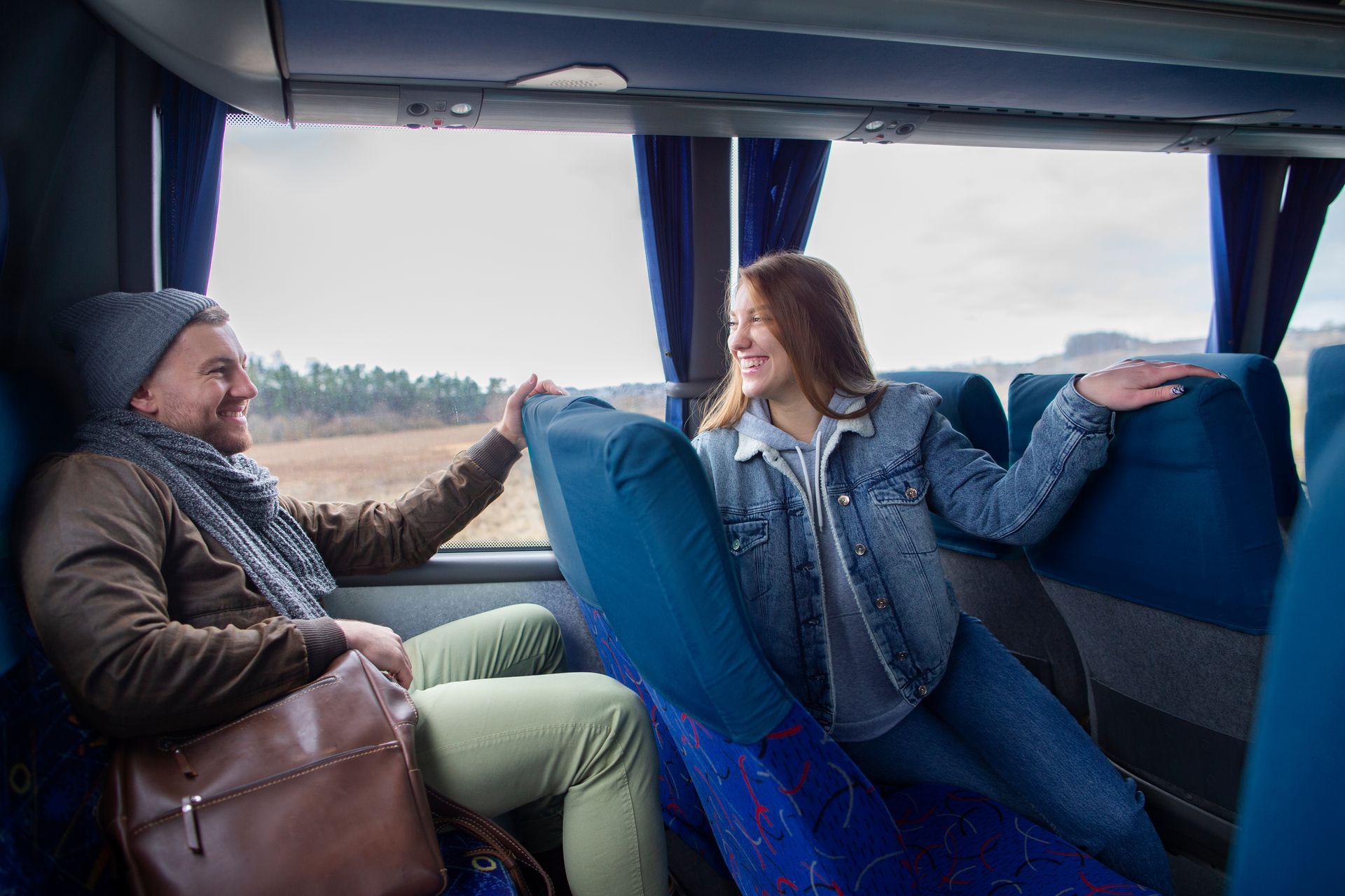 Casal em um ônibus, sorrindo e de mãos dadas, sentado perto da janela. Homem de chapéu e cachecol. Mulher de jaqueta jeans.