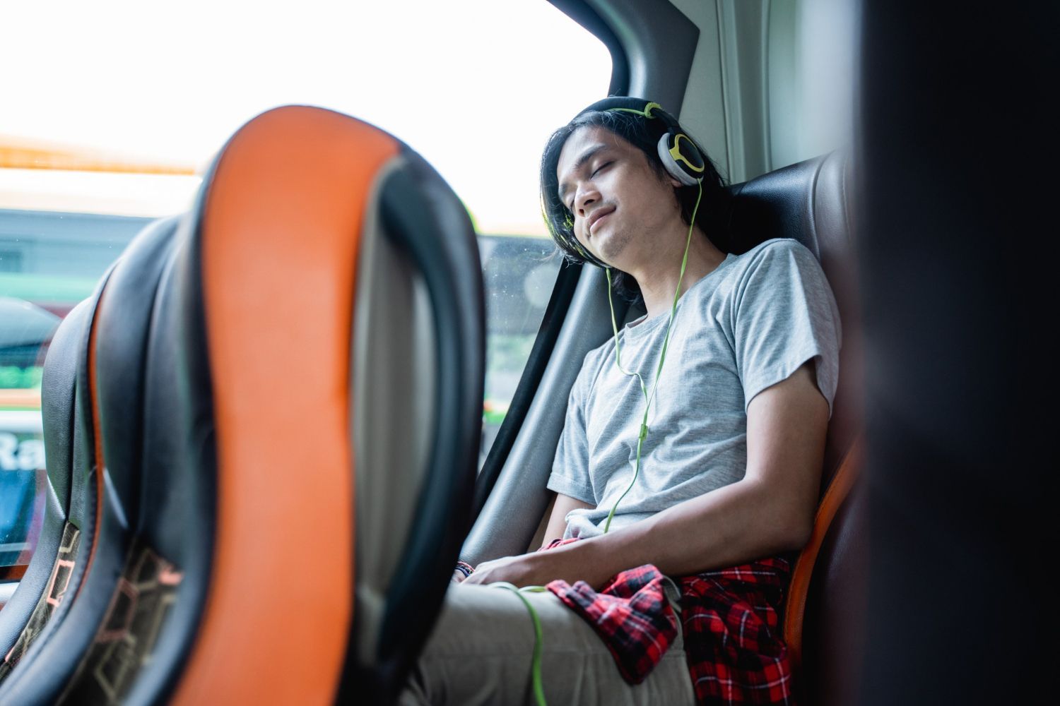 Homem dormindo em um ônibus, usando fones de ouvido, cabeça apoiada na janela, camisa xadrez visível.