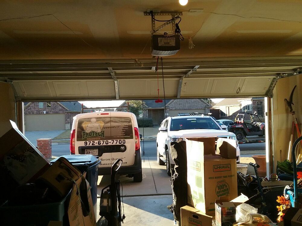 View from inside a garage toward a driveway with a white van, truck, and stacked boxes in the foreground