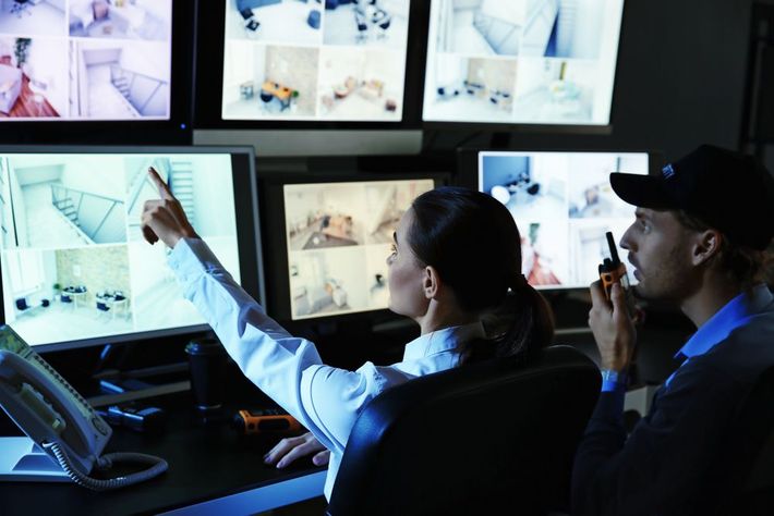 Two operators monitor multiple security camera feeds in a dark surveillance room.