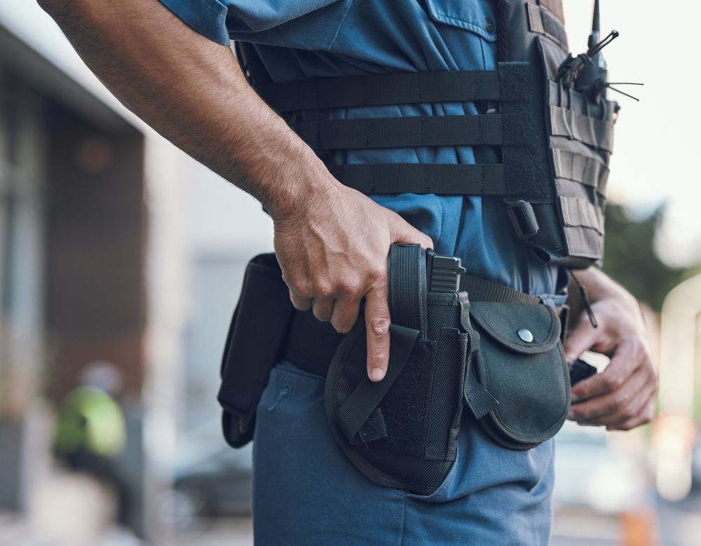 Close-up of a person in a tactical vest holding a black holstered sidearm at their hip