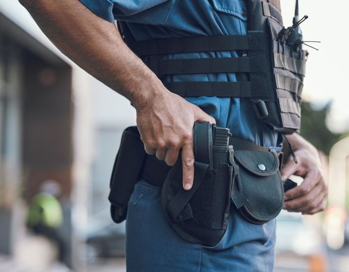 Close-up of a person in a tactical vest holding a black holstered sidearm at their hip