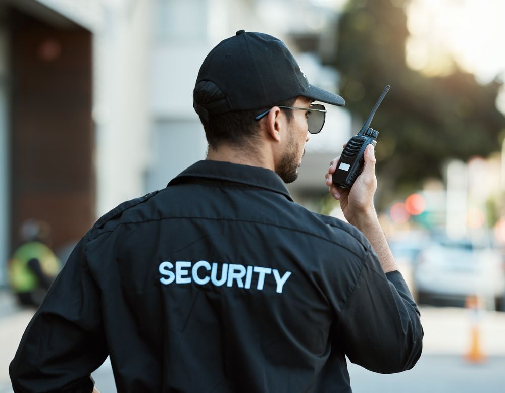 Security guard in black jacket and cap using a walkie-talkie outdoors.