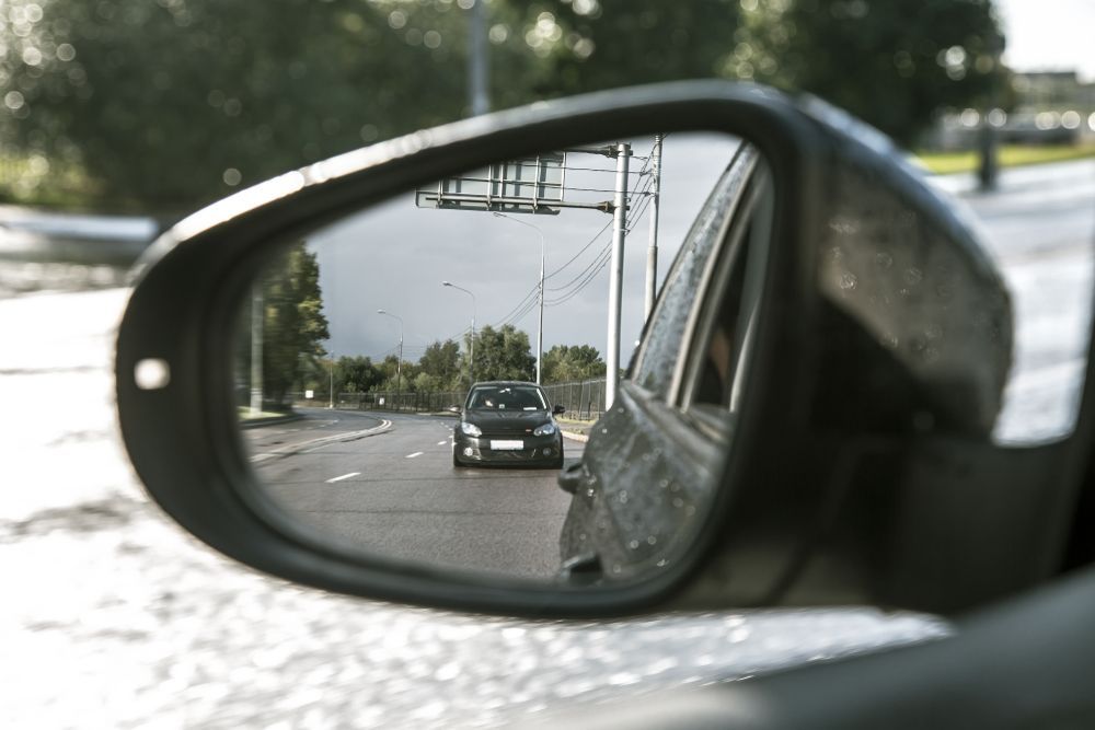 Car seen in side mirror driving behind on a roadside with trees and utility poles.