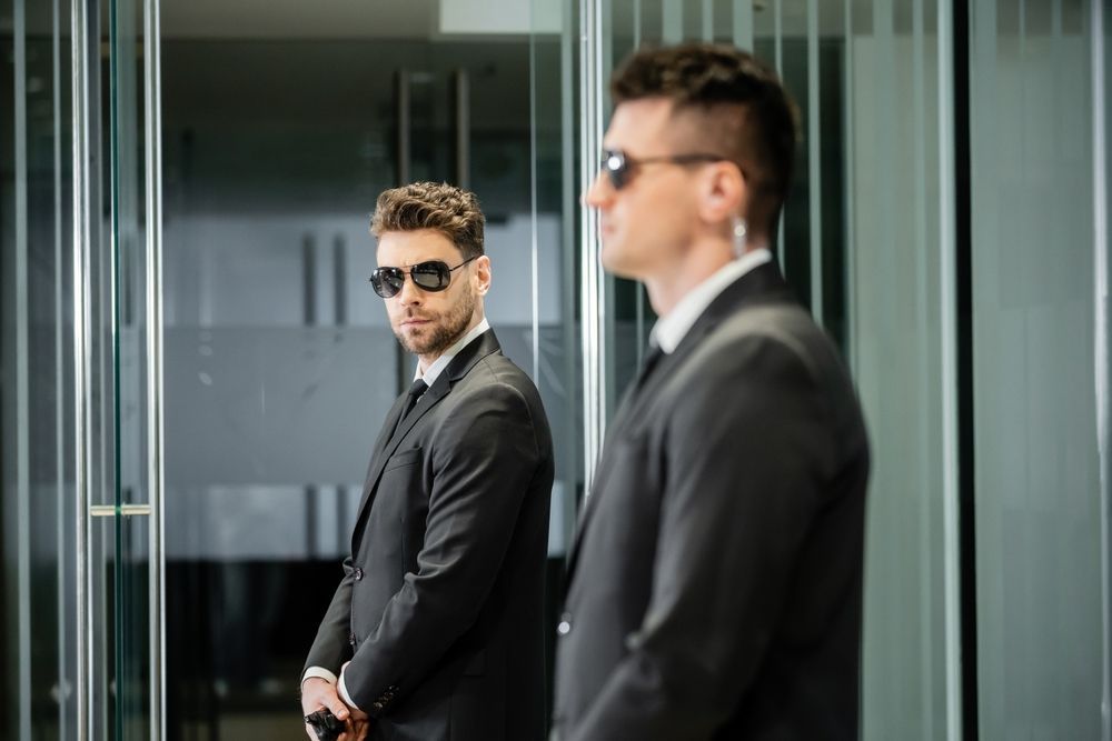 Two men in black suits and sunglasses in a modern glass office hallway