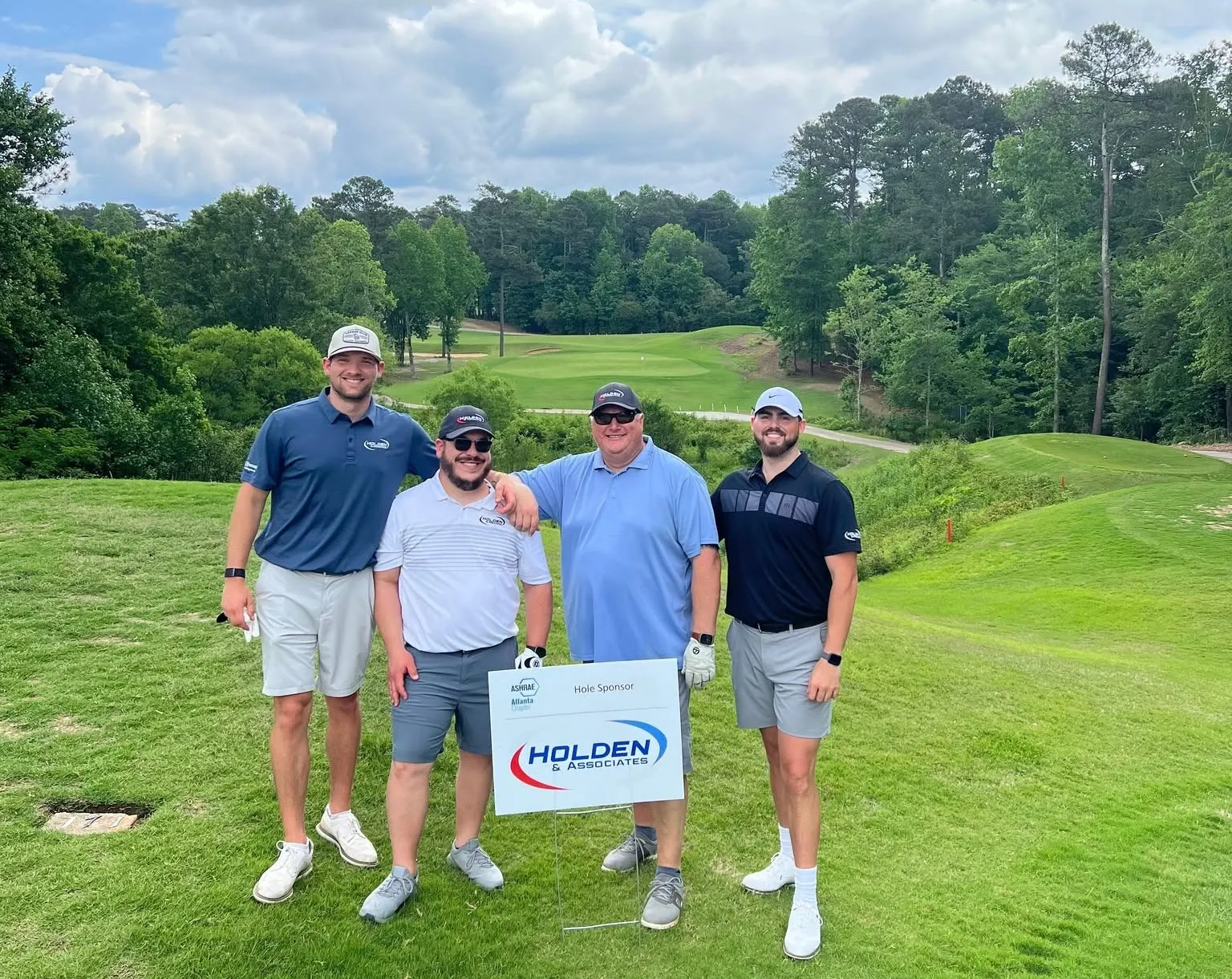 Four golfers holding a sign stand on a green golf course with trees and hills behind them.