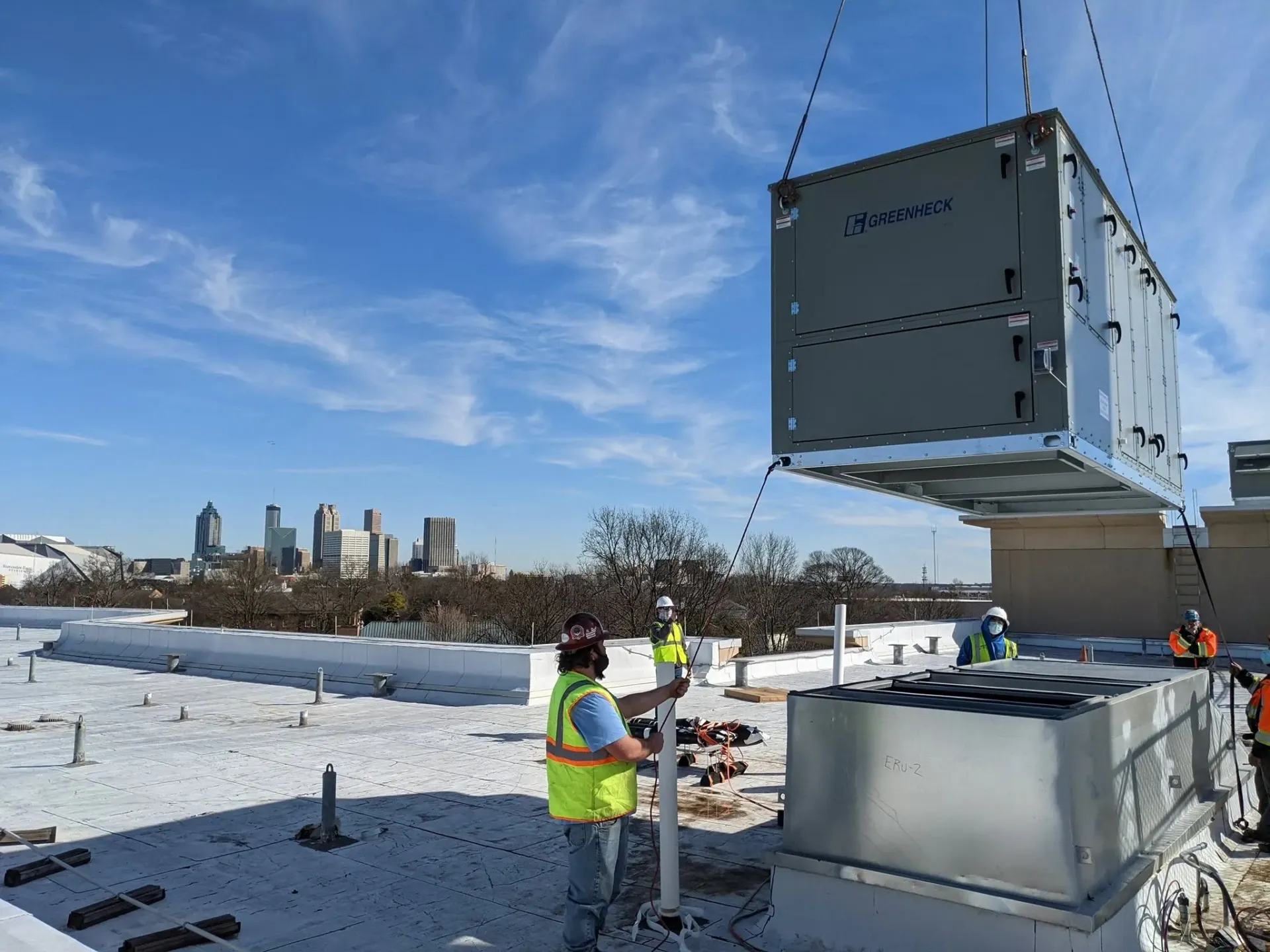 Construction workers lifting a large gray structure on a snowy rooftop with a city skyline in the background