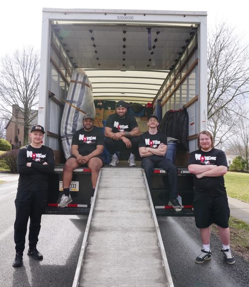 Five Move Me Moving Team Members in front of an open moving truck with a ramp in a parking lot.