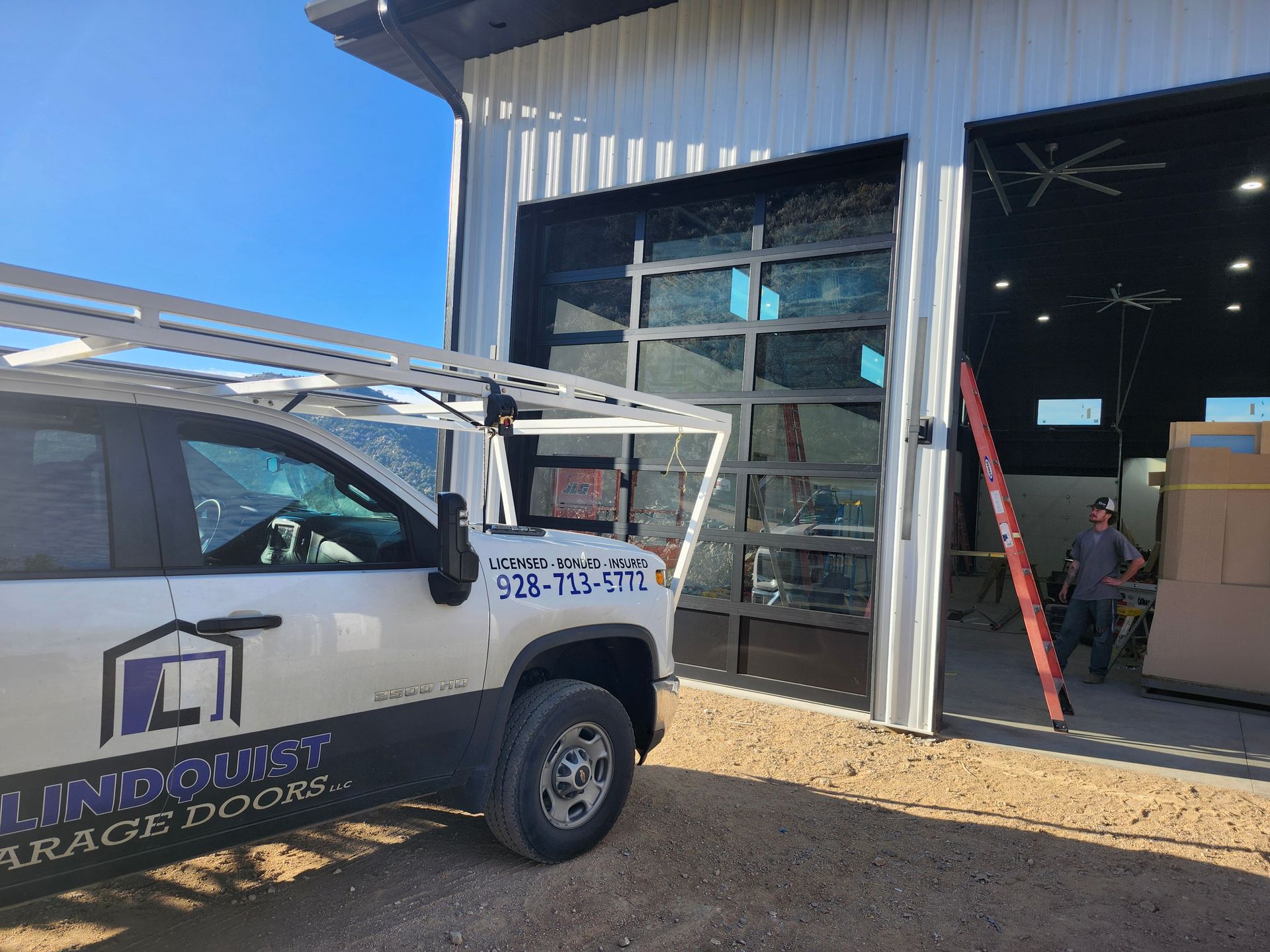 Truck with ladder rack parked outside a building with glass garage door. A person stands inside the open bay door.