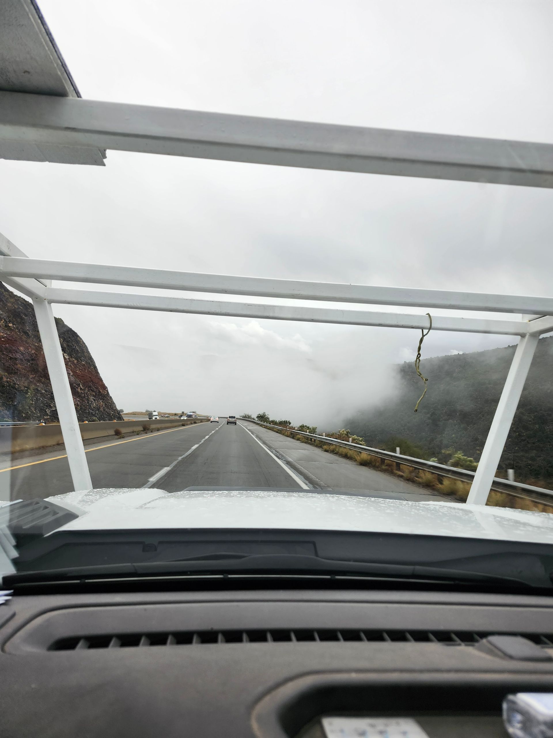 View from a vehicle's windshield on a mountain road with fog and overcast sky.