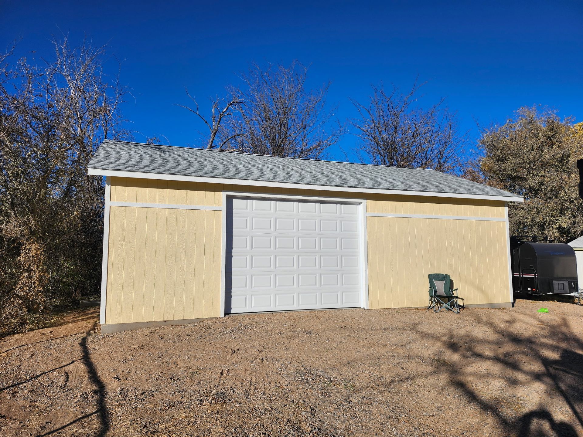 Yellow shed with white door, grey roof, and a chair outside on a gravel lot.
