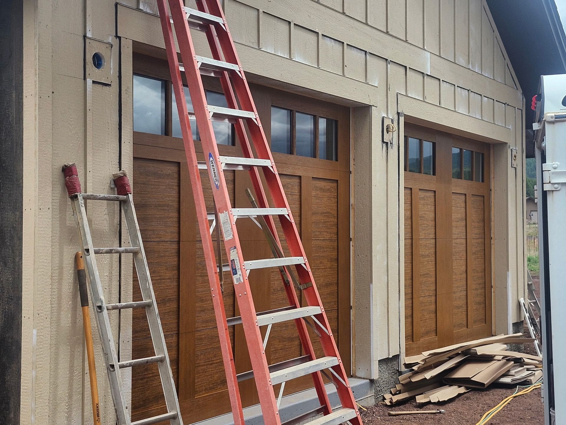Two brown garage doors, tan siding, red and silver ladders, and wood debris outside.