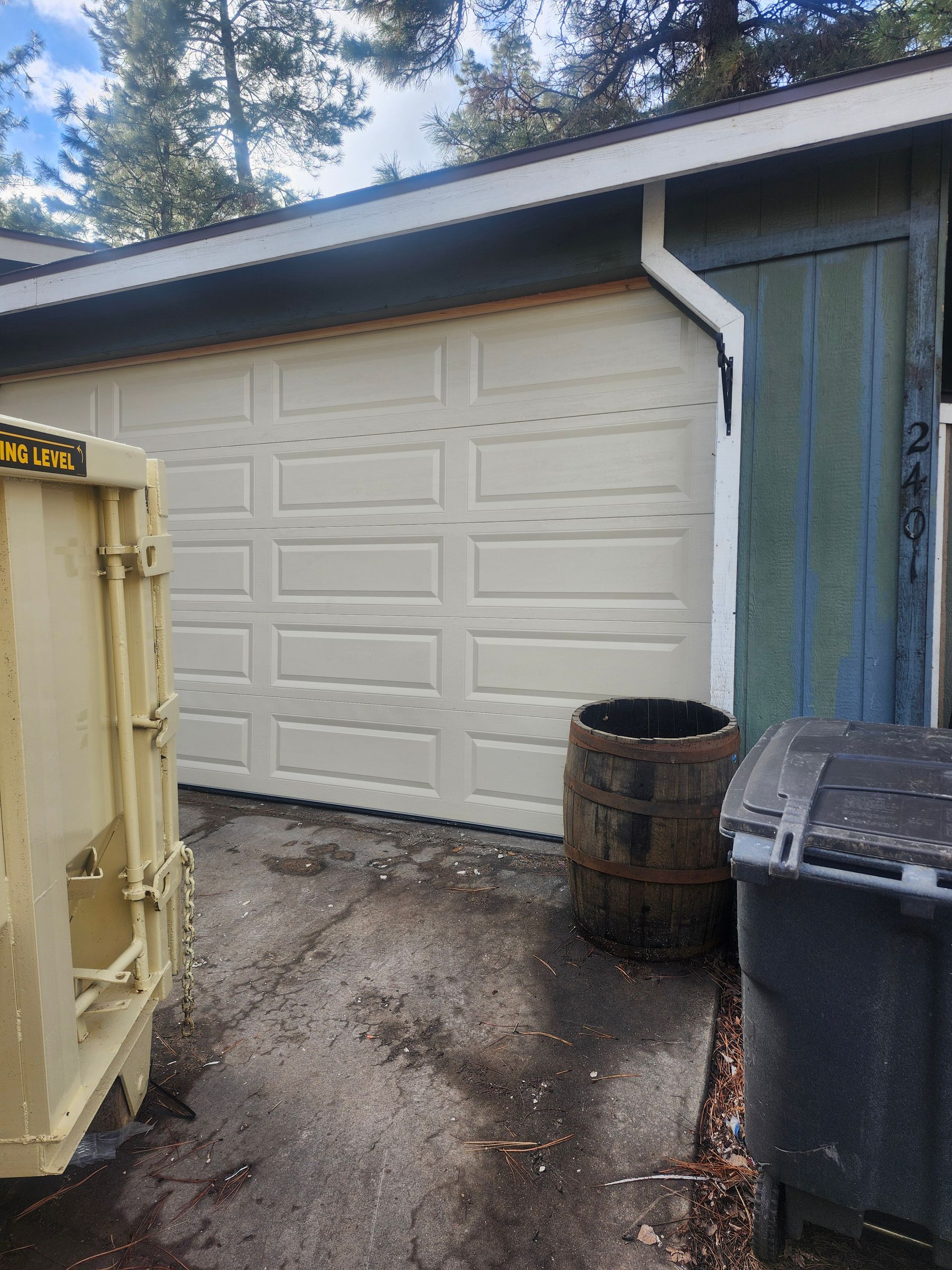 Tan garage door beside blue-green siding. A barrel and trash bins sit near the door.