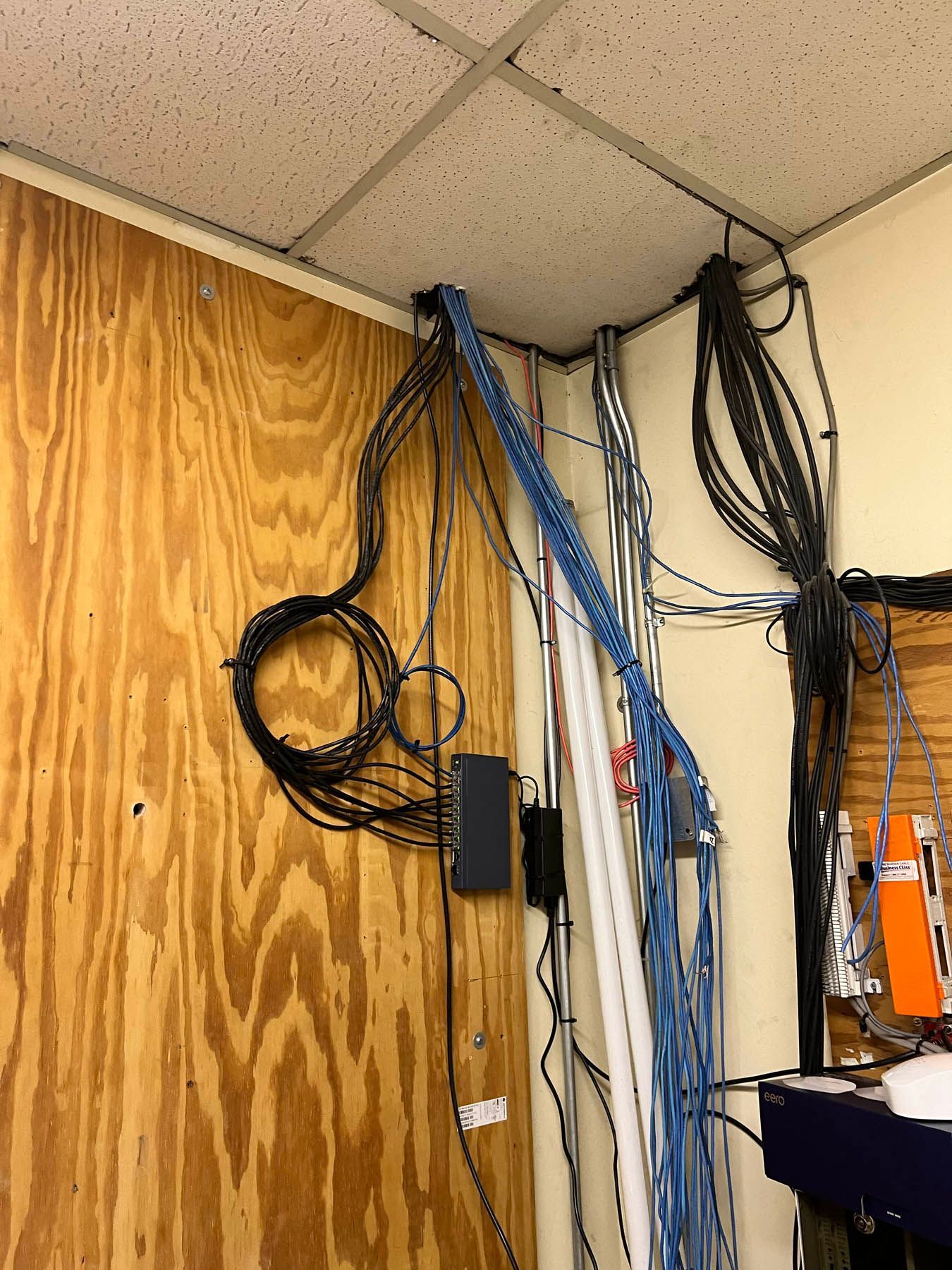 Wall with a chaotic network of black, blue, and white wires, cords, and cables, with a black box attached.
