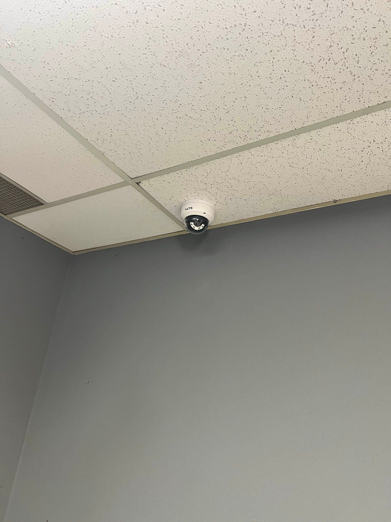 Security camera mounted on a white ceiling with acoustic tiles, above a gray wall.