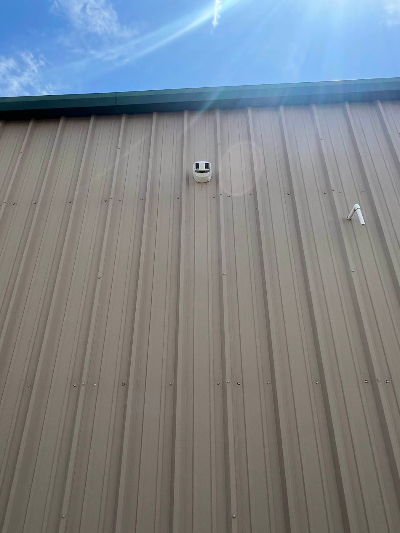 Tan metal building exterior with security camera and pipe against a blue sky.