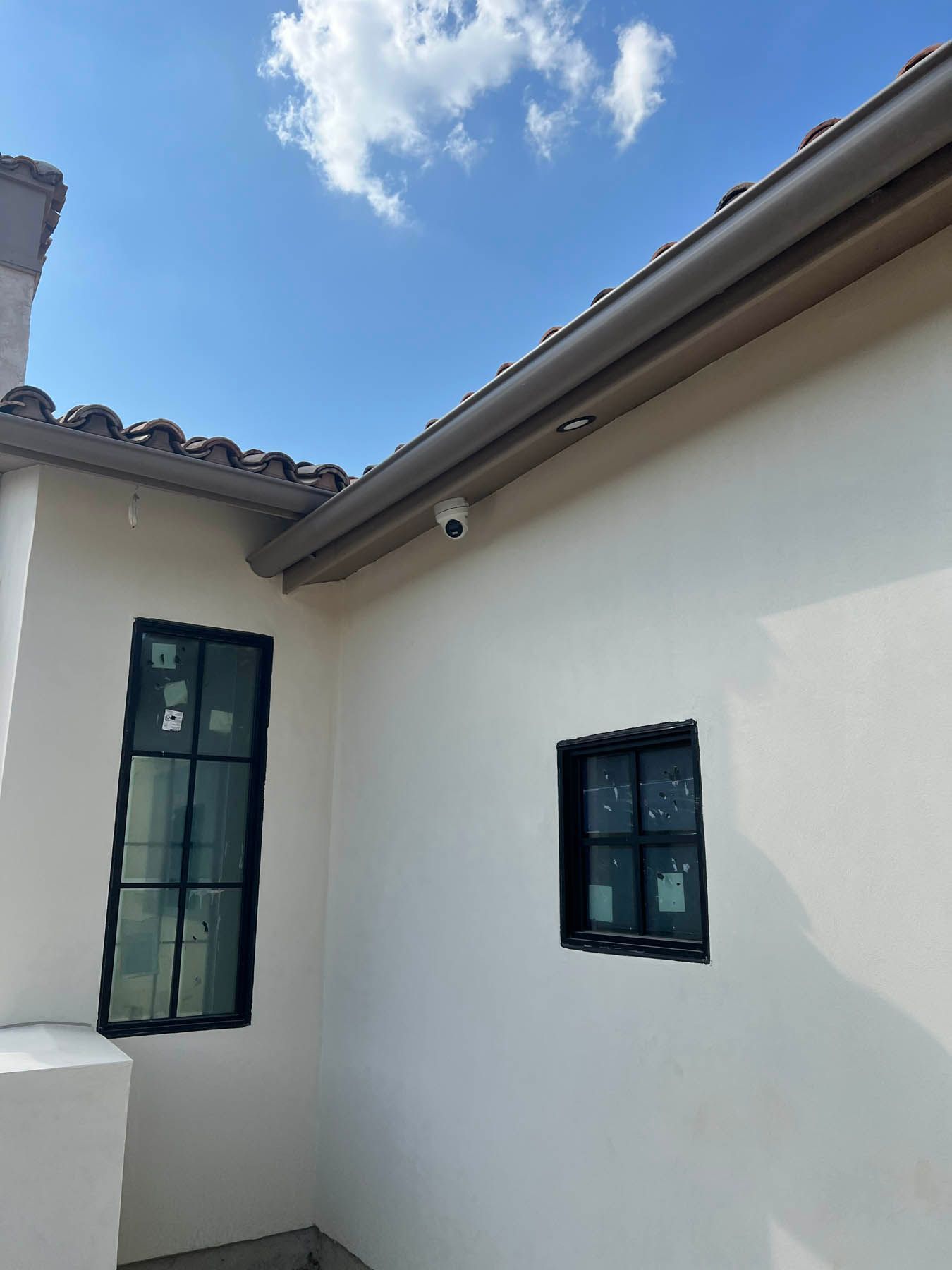 Exterior of a stucco building with a security camera, two windows, brown roof tiles, and a blue sky with clouds.