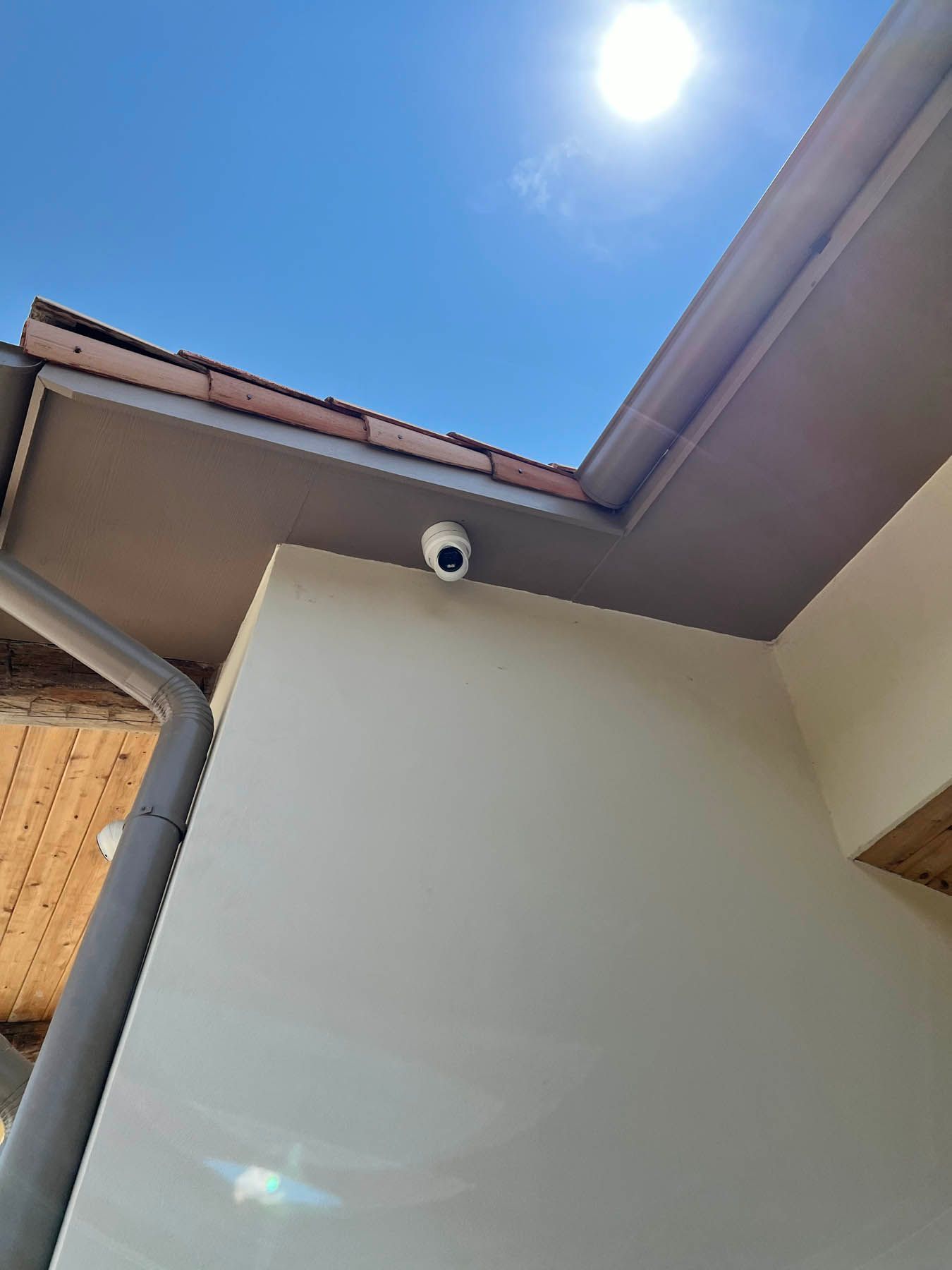 Security camera mounted on a beige building exterior under a light-colored roof, bright sun above.