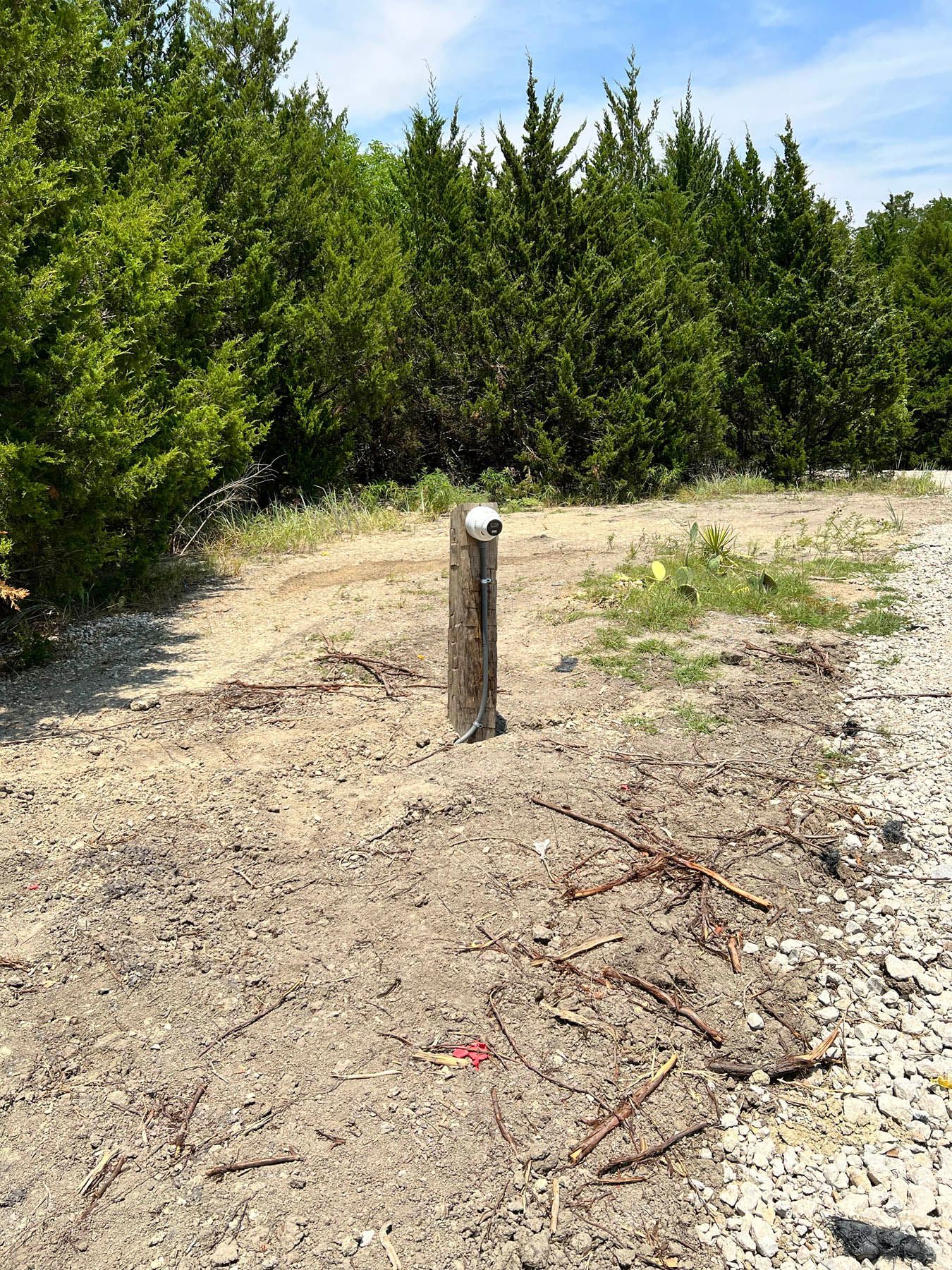 A wooden post with a metal cap stands in a sandy area, trees in the background.