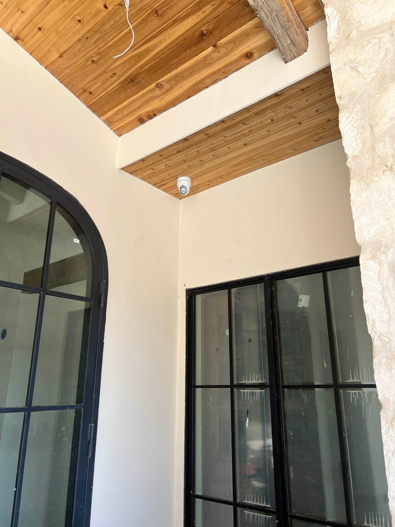 White security camera mounted on a beige wall, with a wood ceiling and black-framed windows.